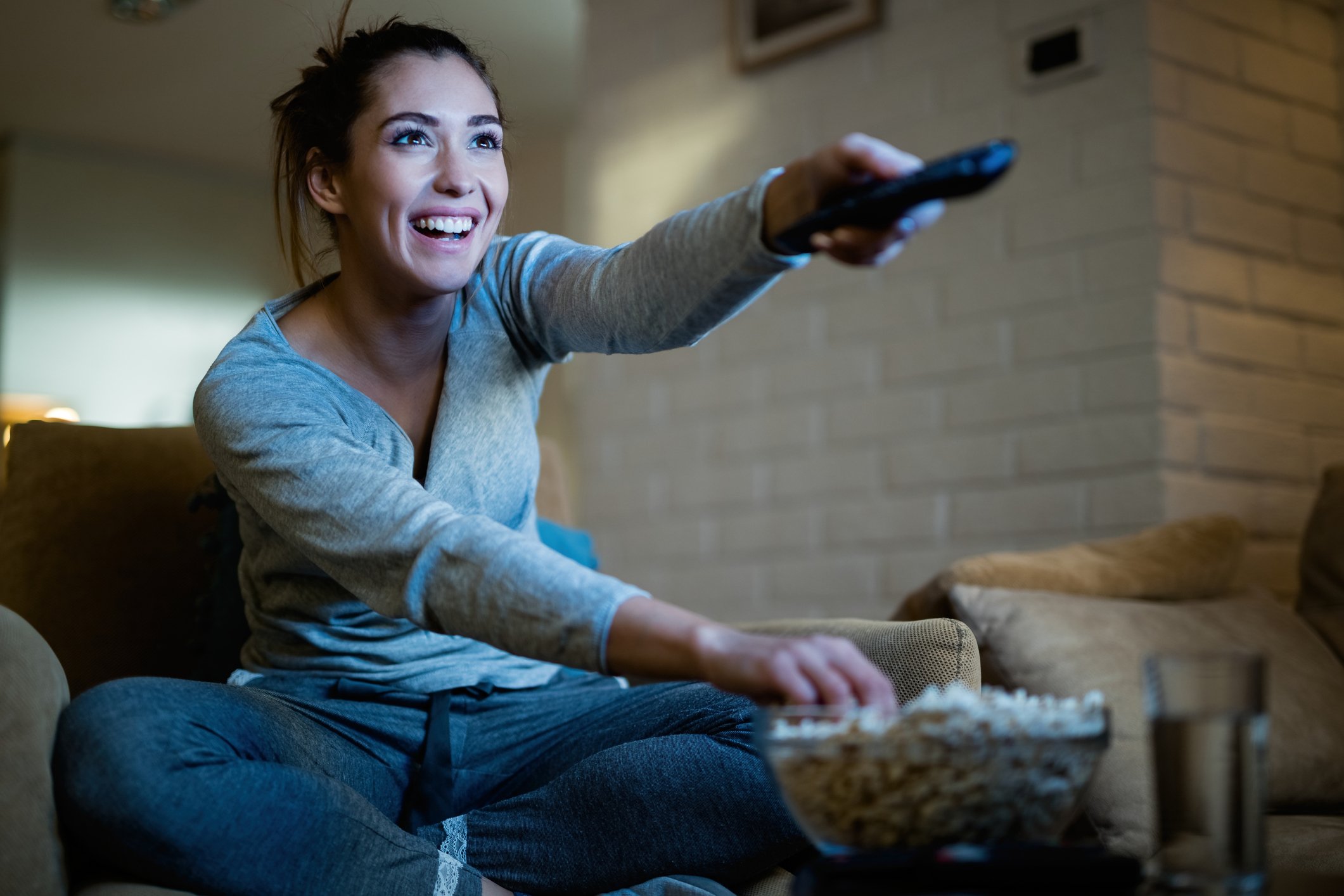 A woman happy to be channel surfing with one hand and grabbing popcorn in the other as she watches TV.