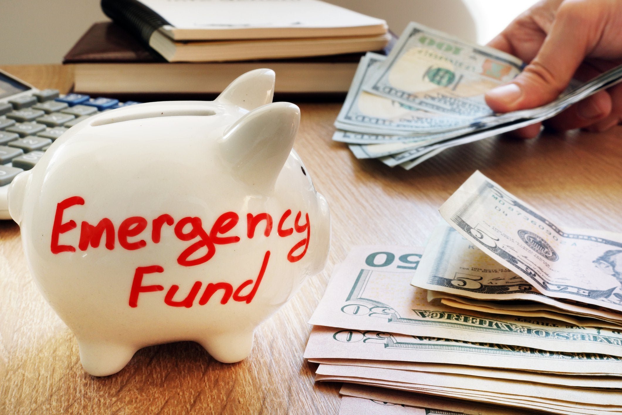 A piggy bank labeled Emergency Fund next to someone counting money at a wooden desk.