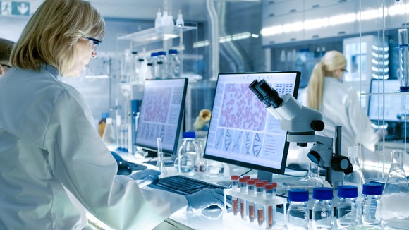A woman at a lab bench with a monitor, microscope, and test tubes.
