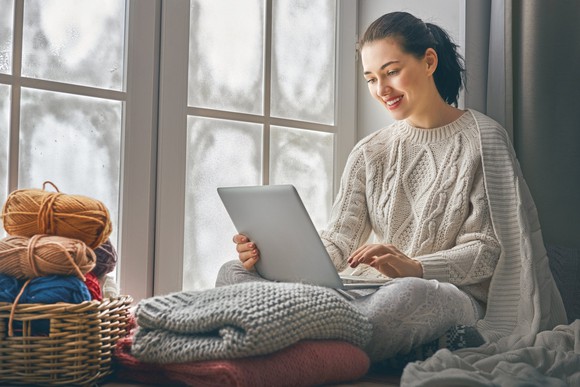 Young woman wearing sweater using laptop next to a window.