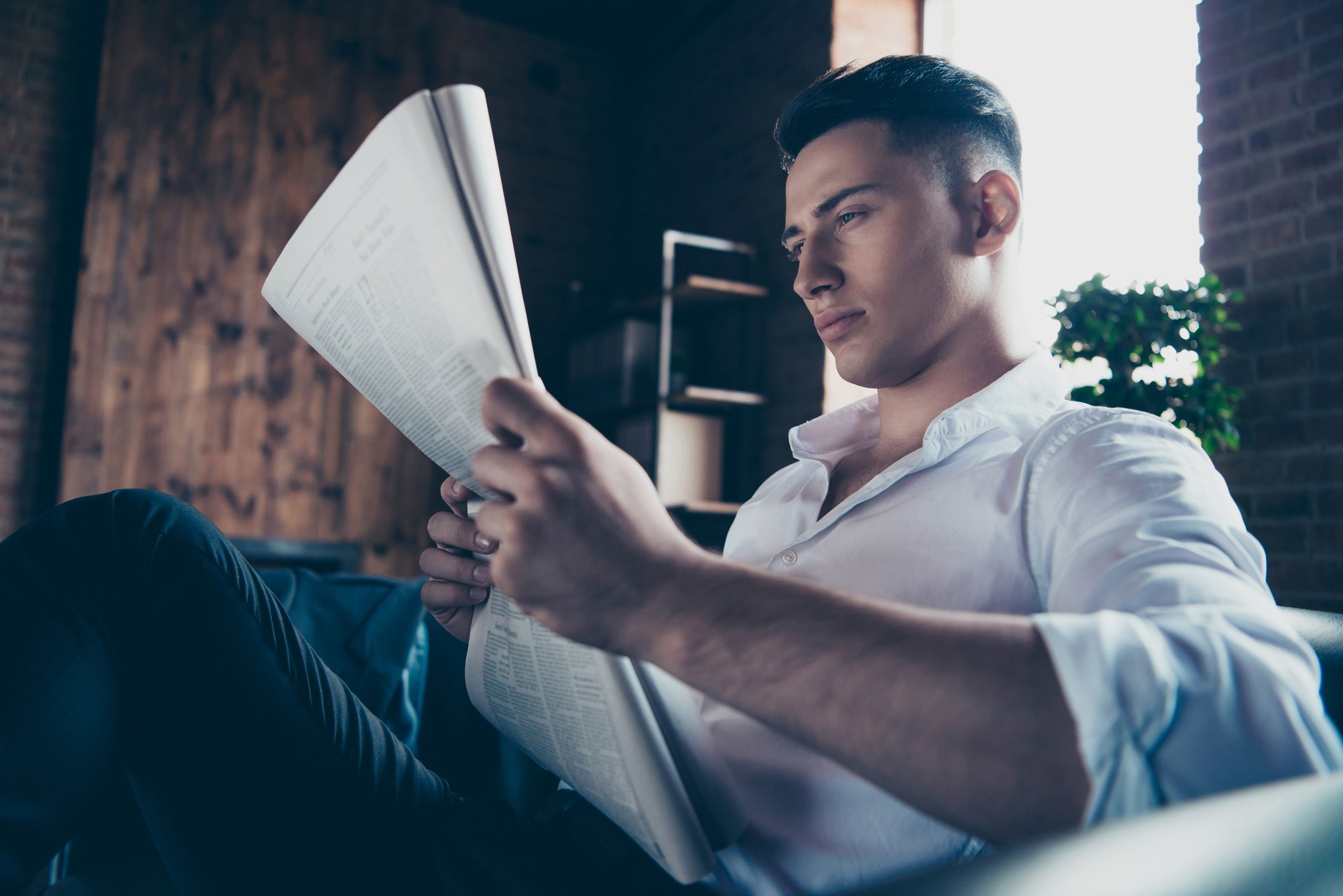 A man sits on a couch, intently reading a newspaper. 