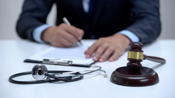 A man at a desk with a gavel and a stethoscope writing on a clipboard.