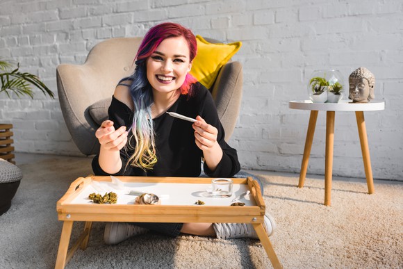A young woman smiles with a joint in her hand in her living room. 