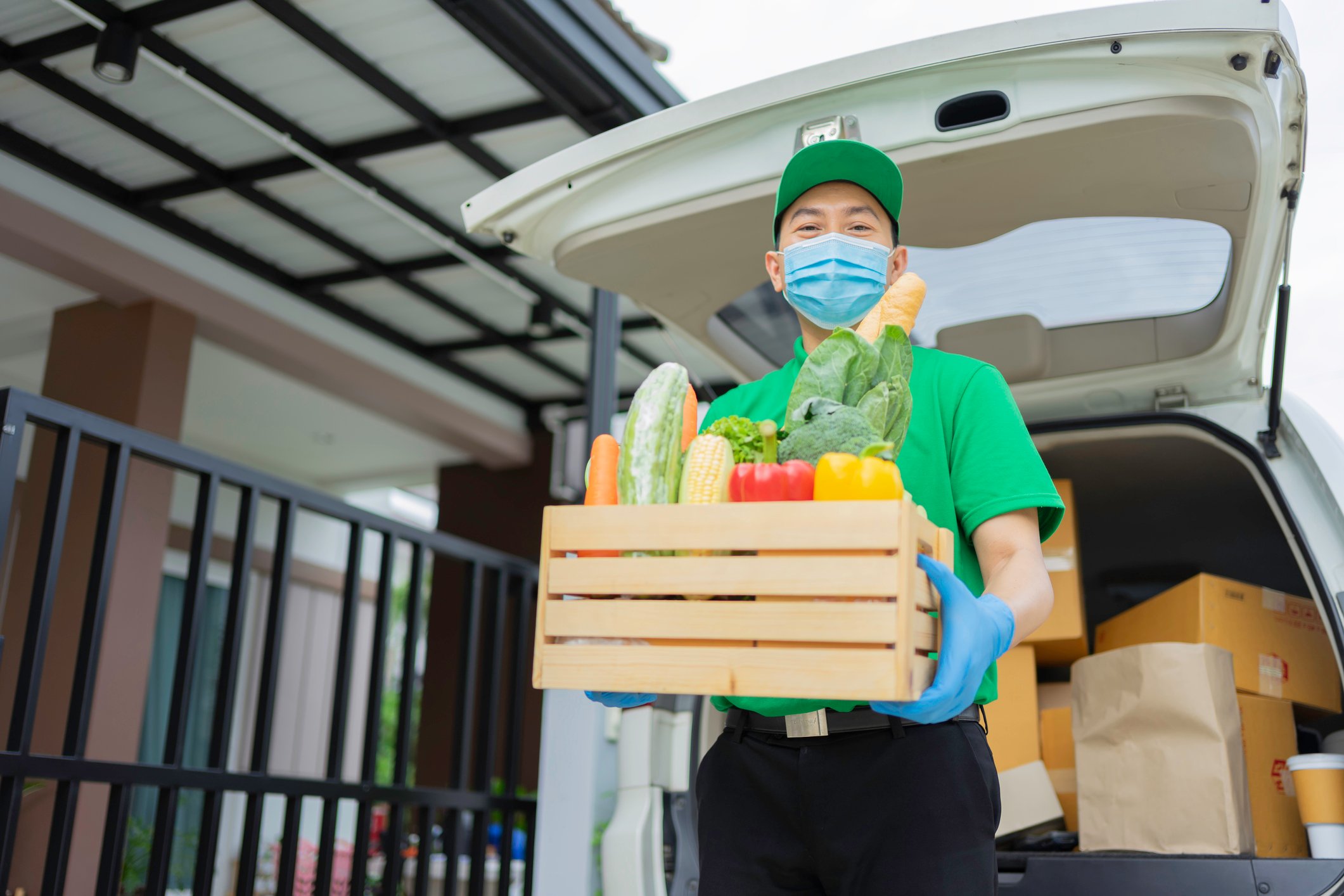 A masked delivery man holds a crate of vegetables outside his delivery car. 