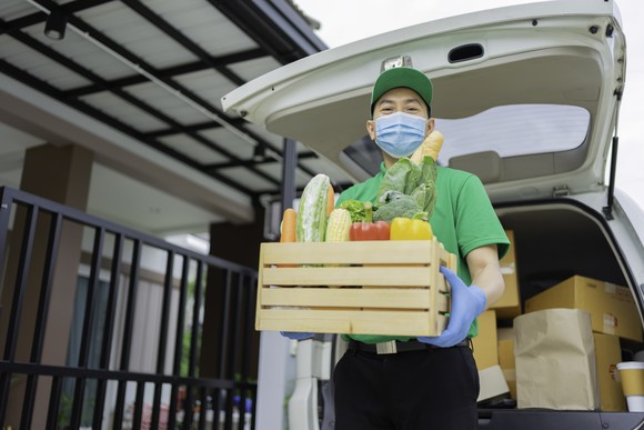 A masked delivery man holds a crate of vegetables outside his delivery car. 