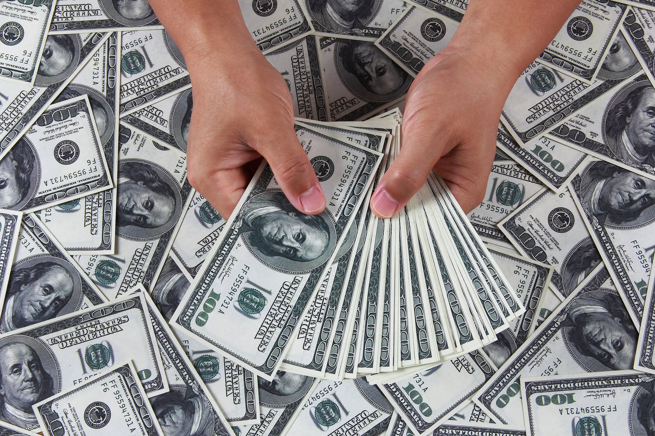 Hands holding $100 bills fanned out over a pile of $100 bills