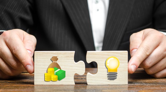 Man wearing a business coat pushing two jigsaw puzzle pieces together, one featuring images of a money bag, cash, and coins, and the other showing a light bulb.