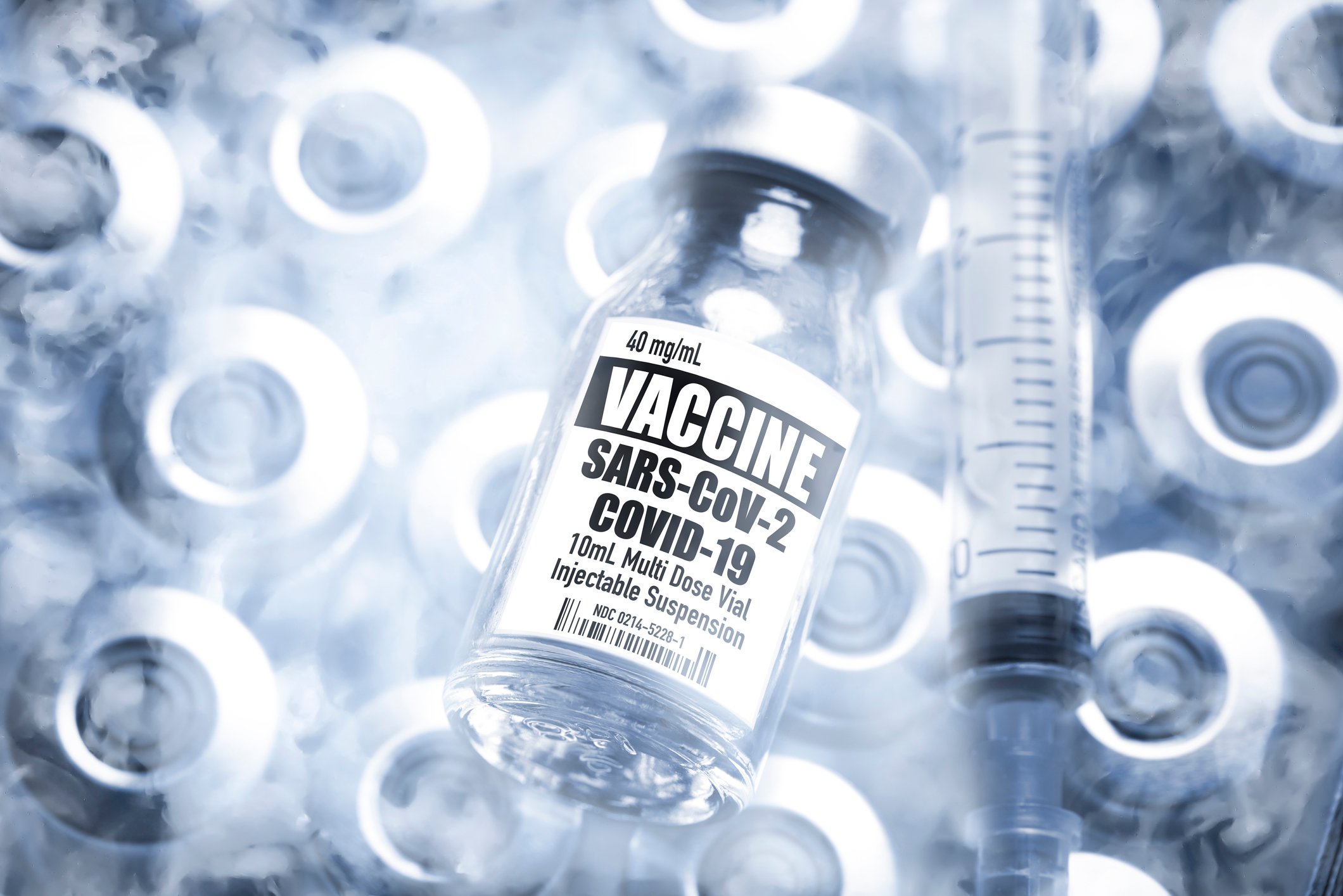 A syringe and a vial labeled Vaccine SARS-CoV-2 COVID-19, in a shipping container with other vials on dry ice