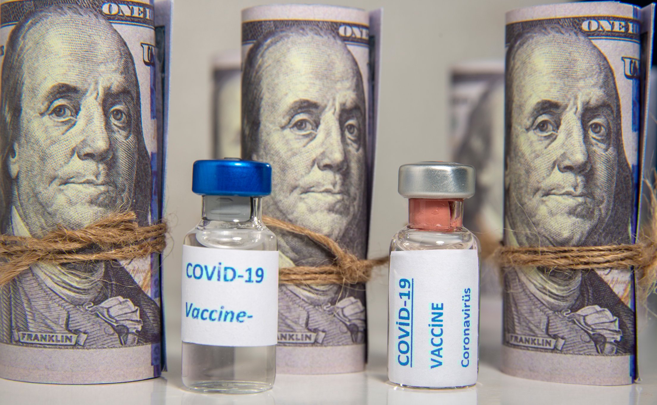 Vials labeled COVID-19 Vaccine, in front of a backdrop of rolled-up $100 bills