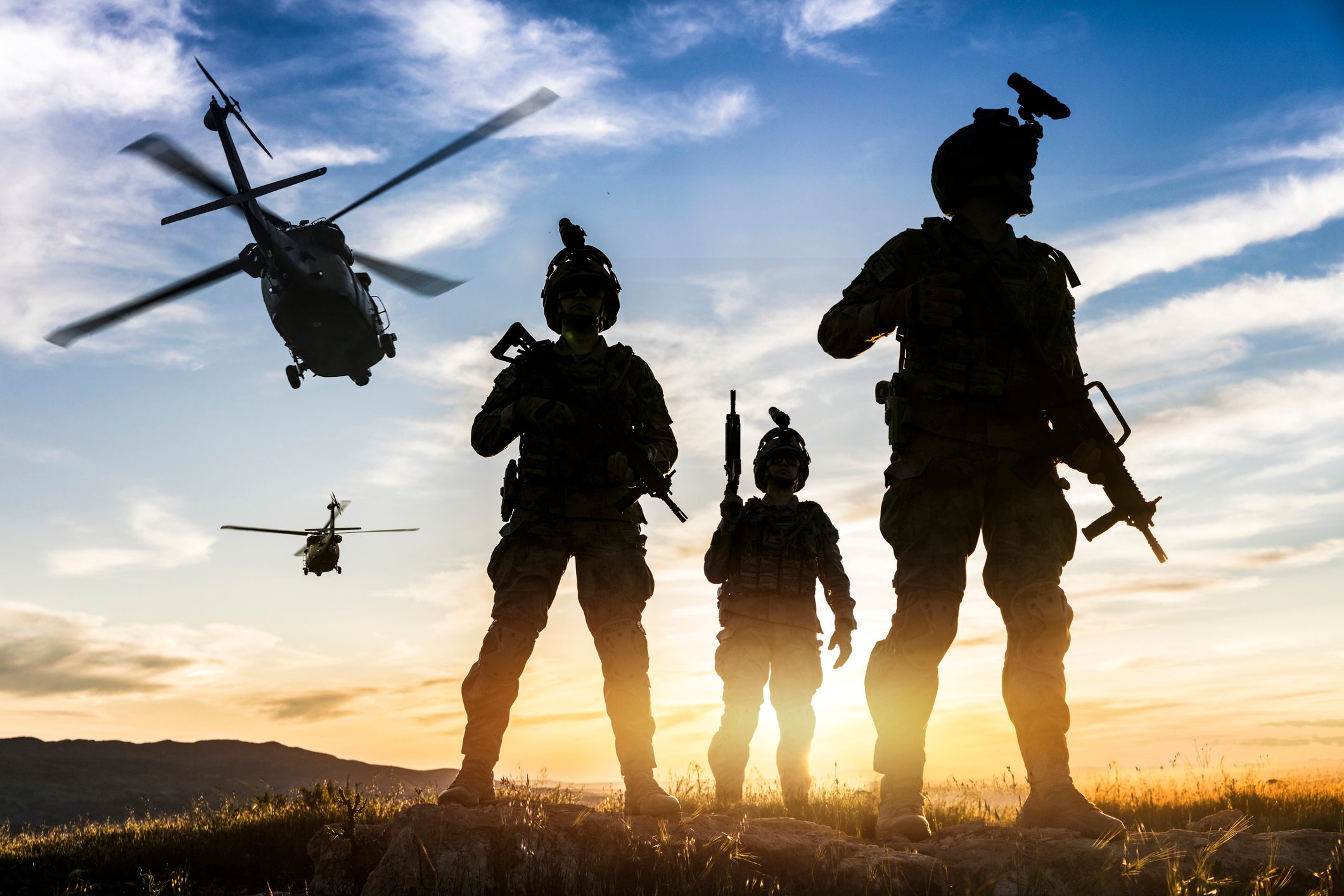 Silhouettes of departing helicopters and three weapons-bearing soldiers standing together. 