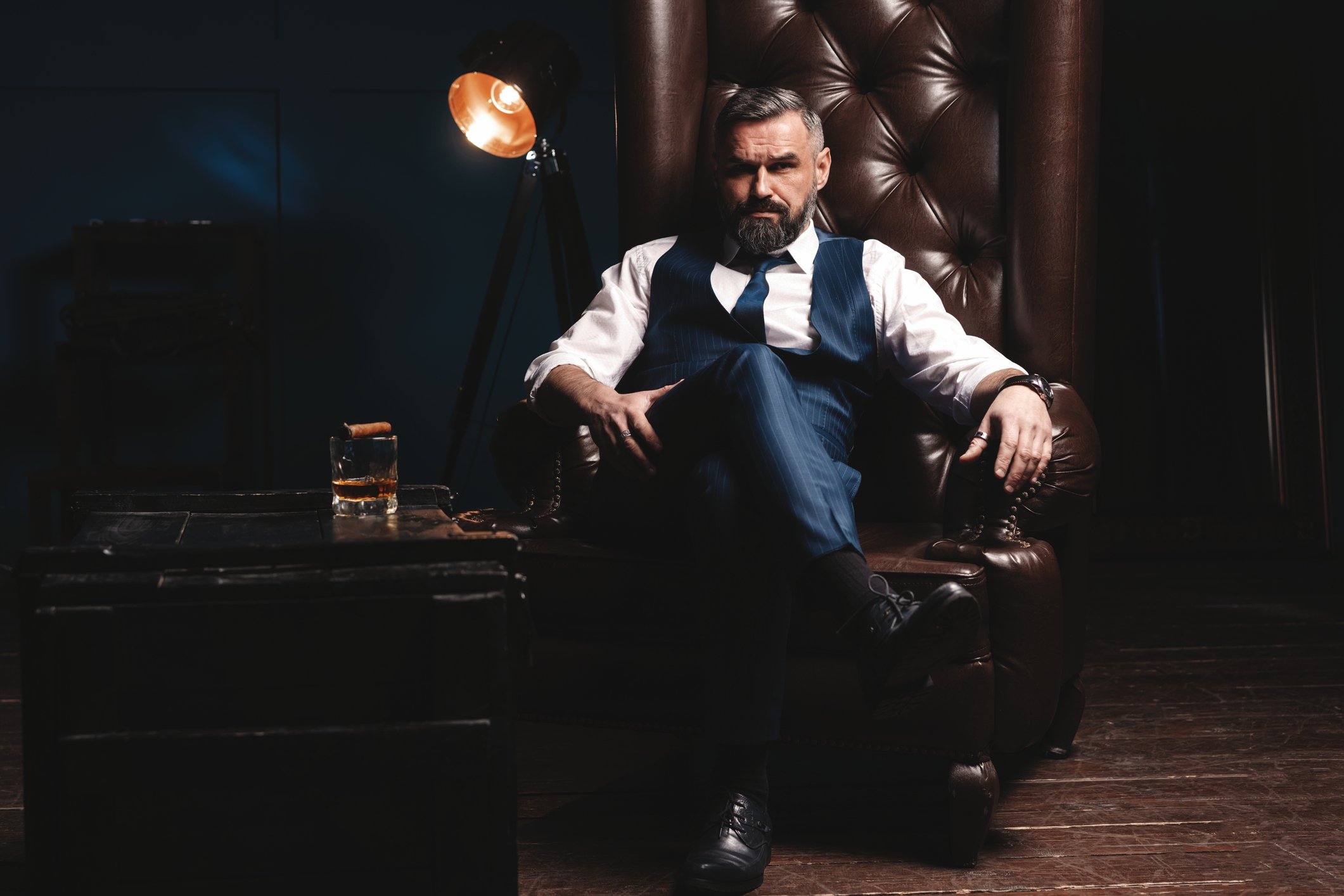 bearded man in oxford shirt, tie, vest, and slacks sitting in leather armchair next to table with glass of whiskey on top