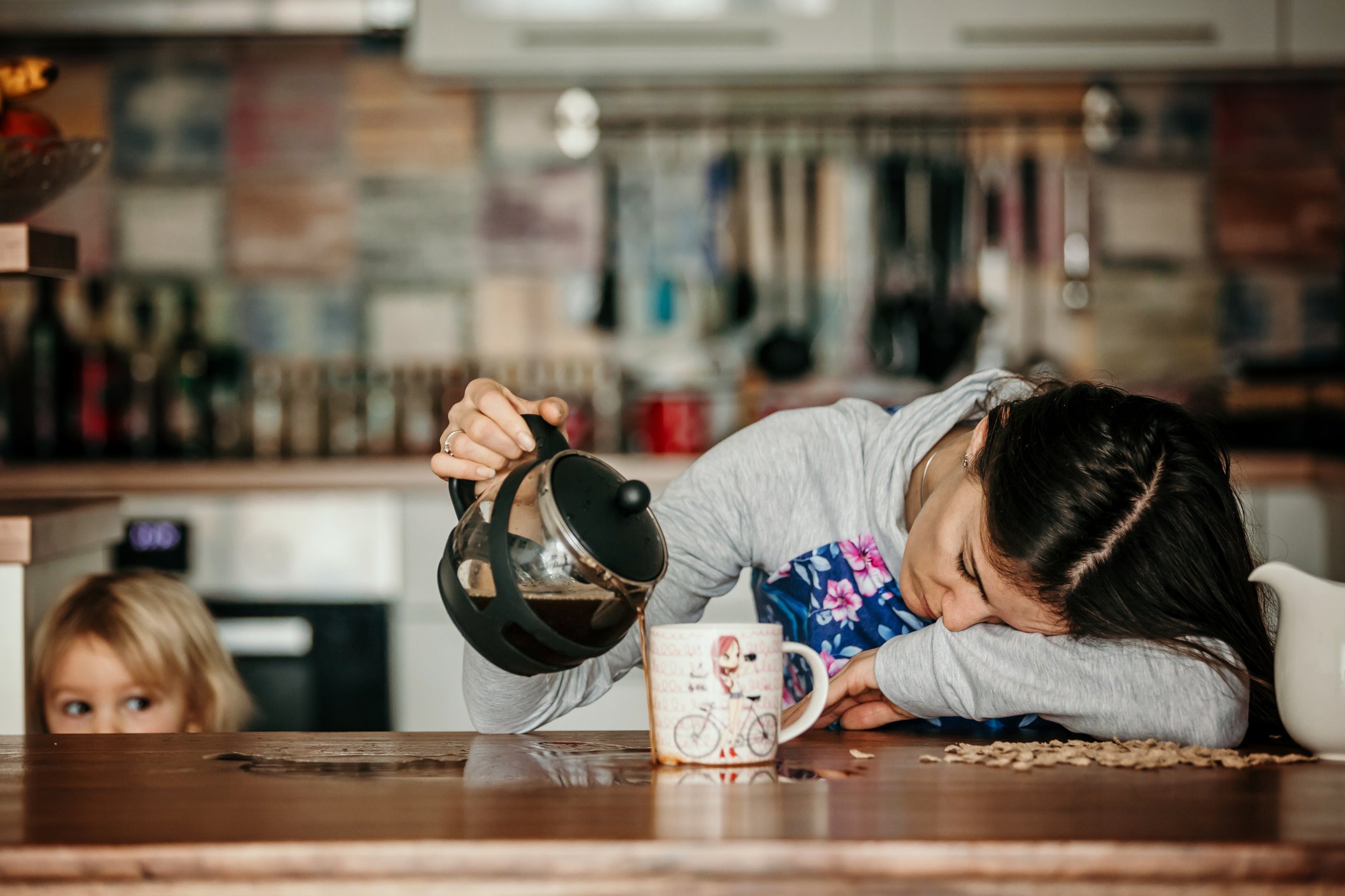 A parent falls asleep on  a counter while pouring coffee in a mug. 