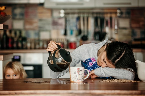 A parent falls asleep on  a counter while pouring coffee in a mug. 