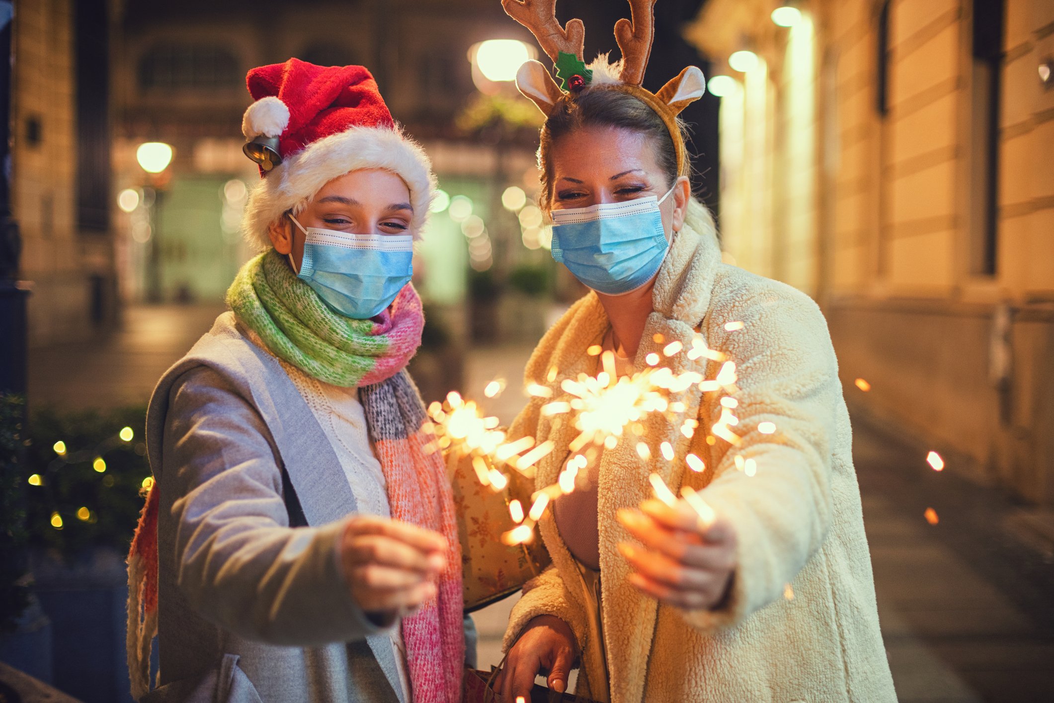 Two women in masks and Christmas headwear, holding sparklers