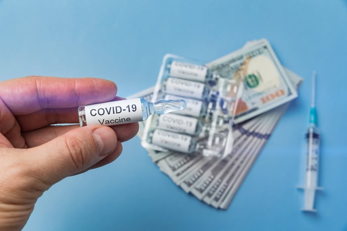 A researcher's hand holds a dose of coronavirus vaccine with $100 bills and a syringe in the background.