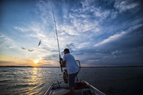 A fisherman on a small boat catching a fish from a lake at sunrise or sunset. 