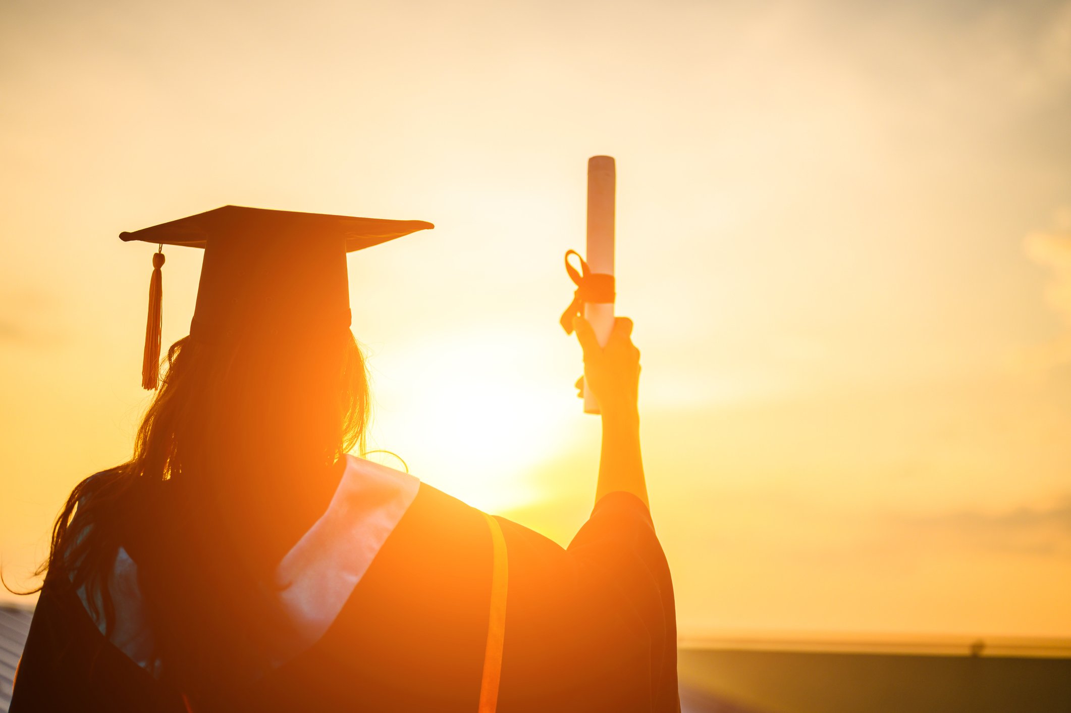 A college graduate in cap and gown holding up a diploma, staring out at the sunset. 