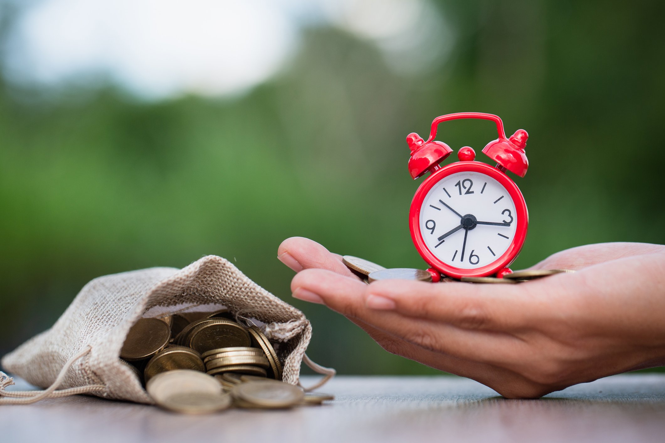 A hand holding a clock and coins alongside a bag of coins.