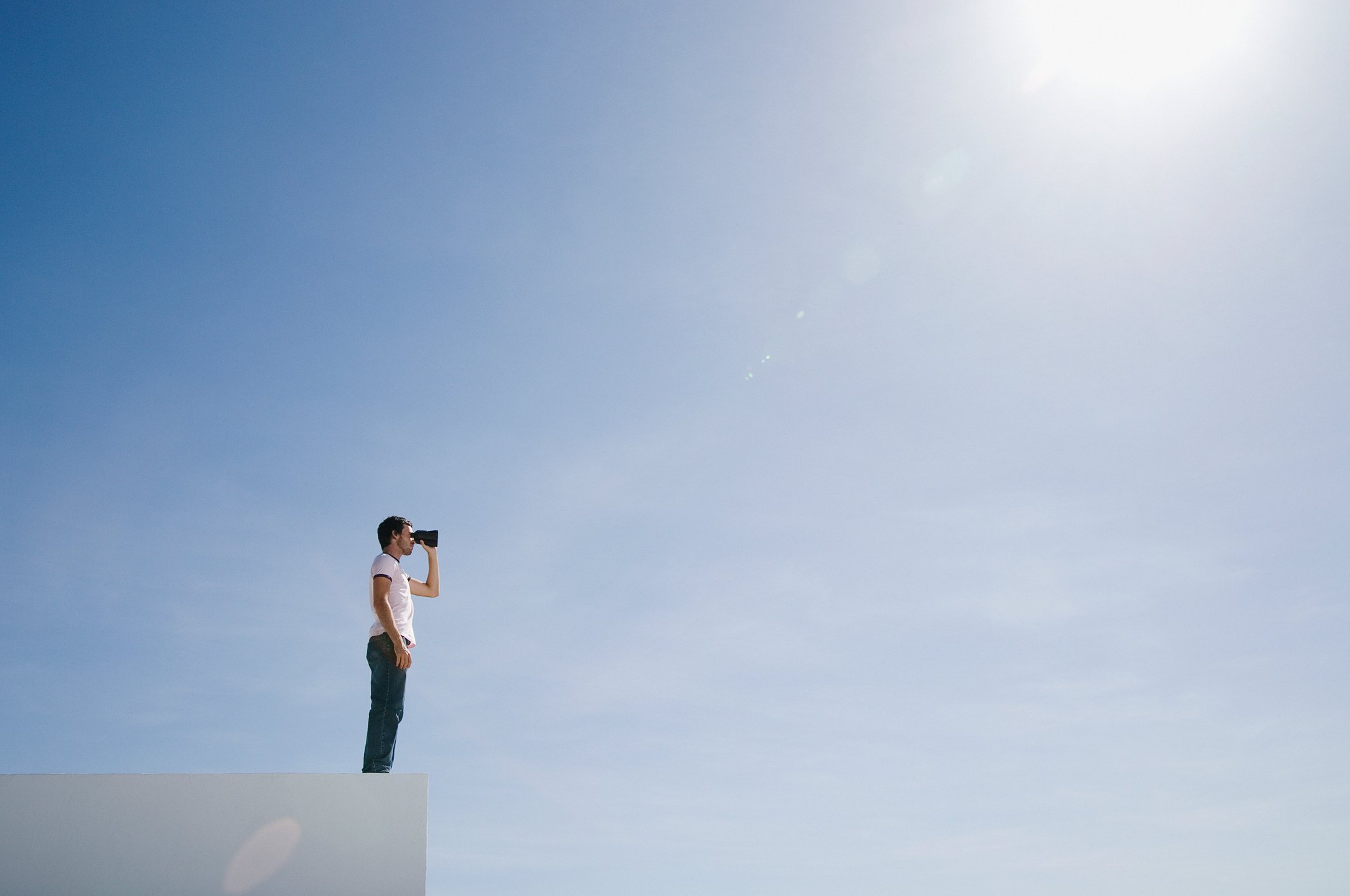 Man looking through binoculars under a blue, sunny sky.