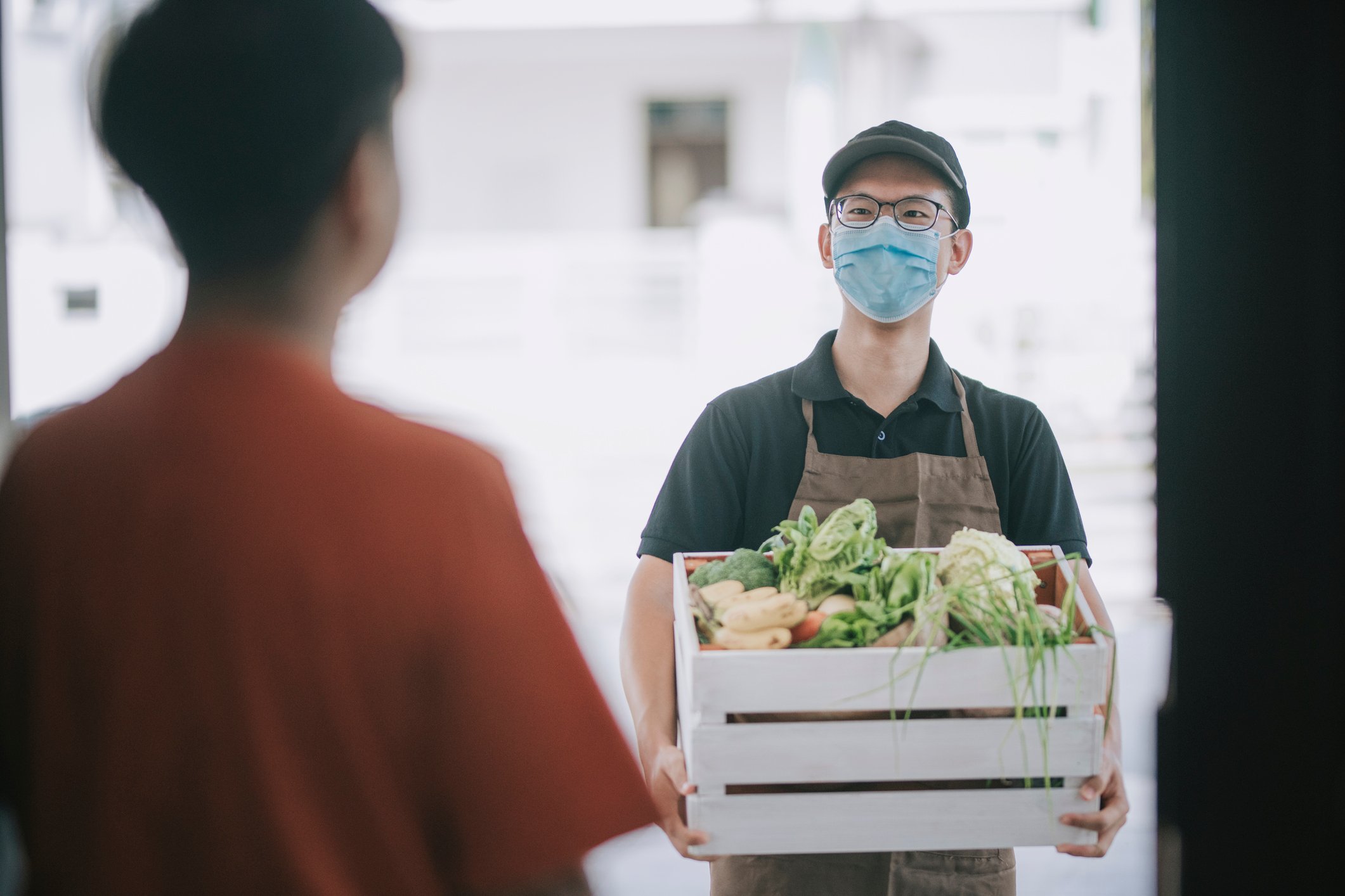 A masked delivery  man stands outside an open front door with a crate of vegetables. 