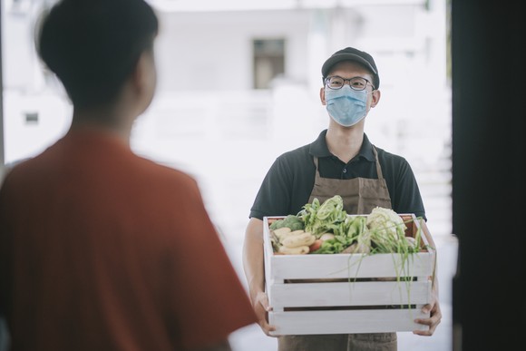 A masked delivery  man stands outside an open front door with a crate of vegetables. 