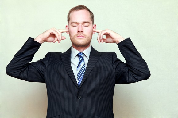 A man in a suit with his eyes closed and fingers in his ears ignoring anything around him.