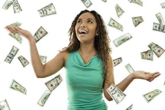 A smiling person stands under a shower of falling U.S. paper currency. 