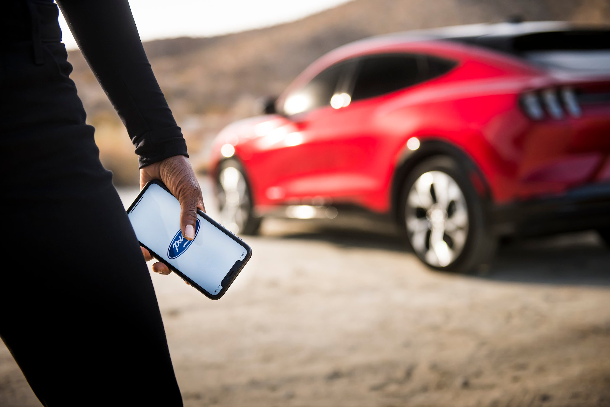 A woman holding a cell phone with a Ford logo faces a red Ford Mustang Mach-E in the background. 