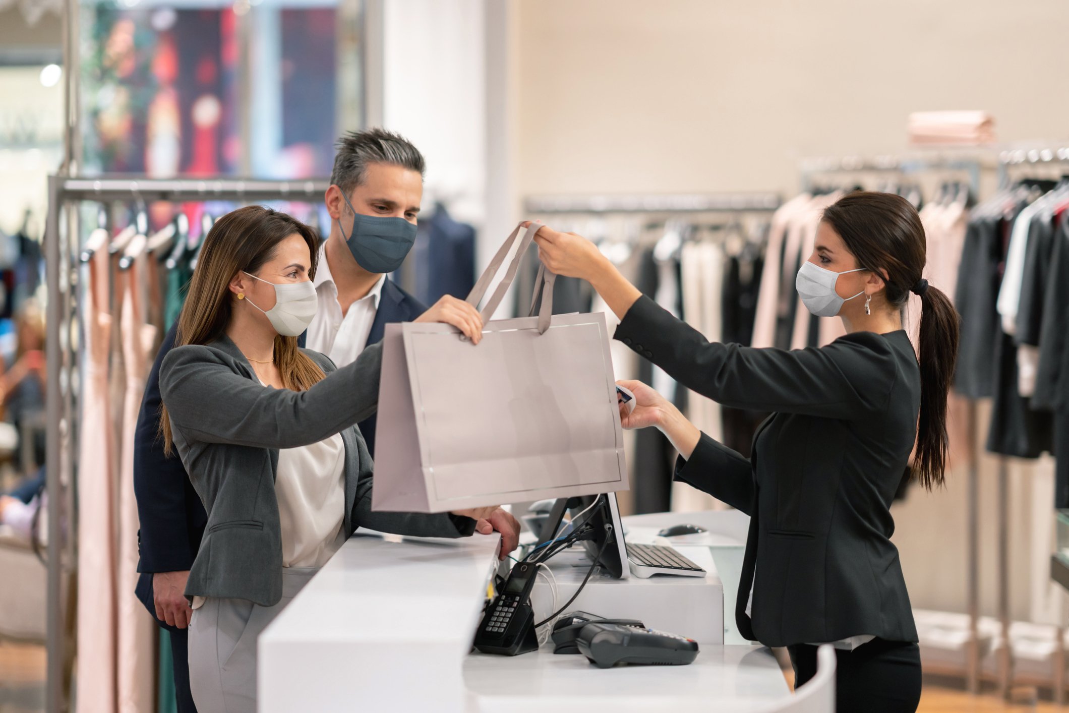 A couple fashion shopping at an apparel retailer, checking out with both shoppers and clerk wearing masks.