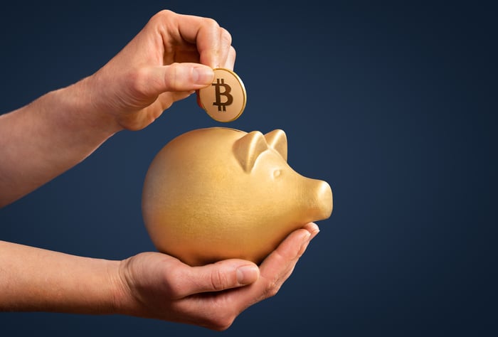 A person holds a golden piggy bank, placing a coin with the bitcoin logo into the bank.