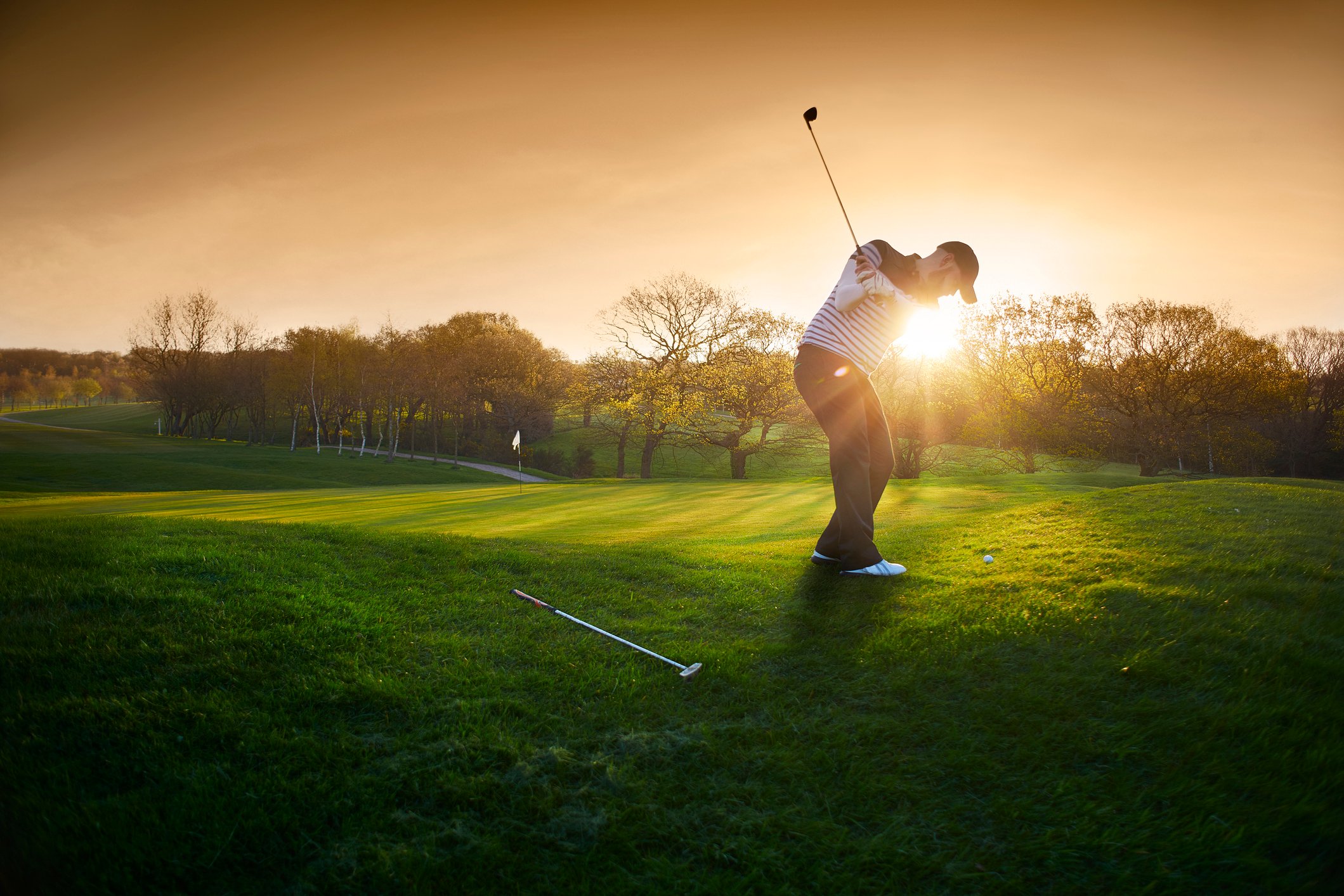 A golfer swings a club near a green as the sun sets. 
