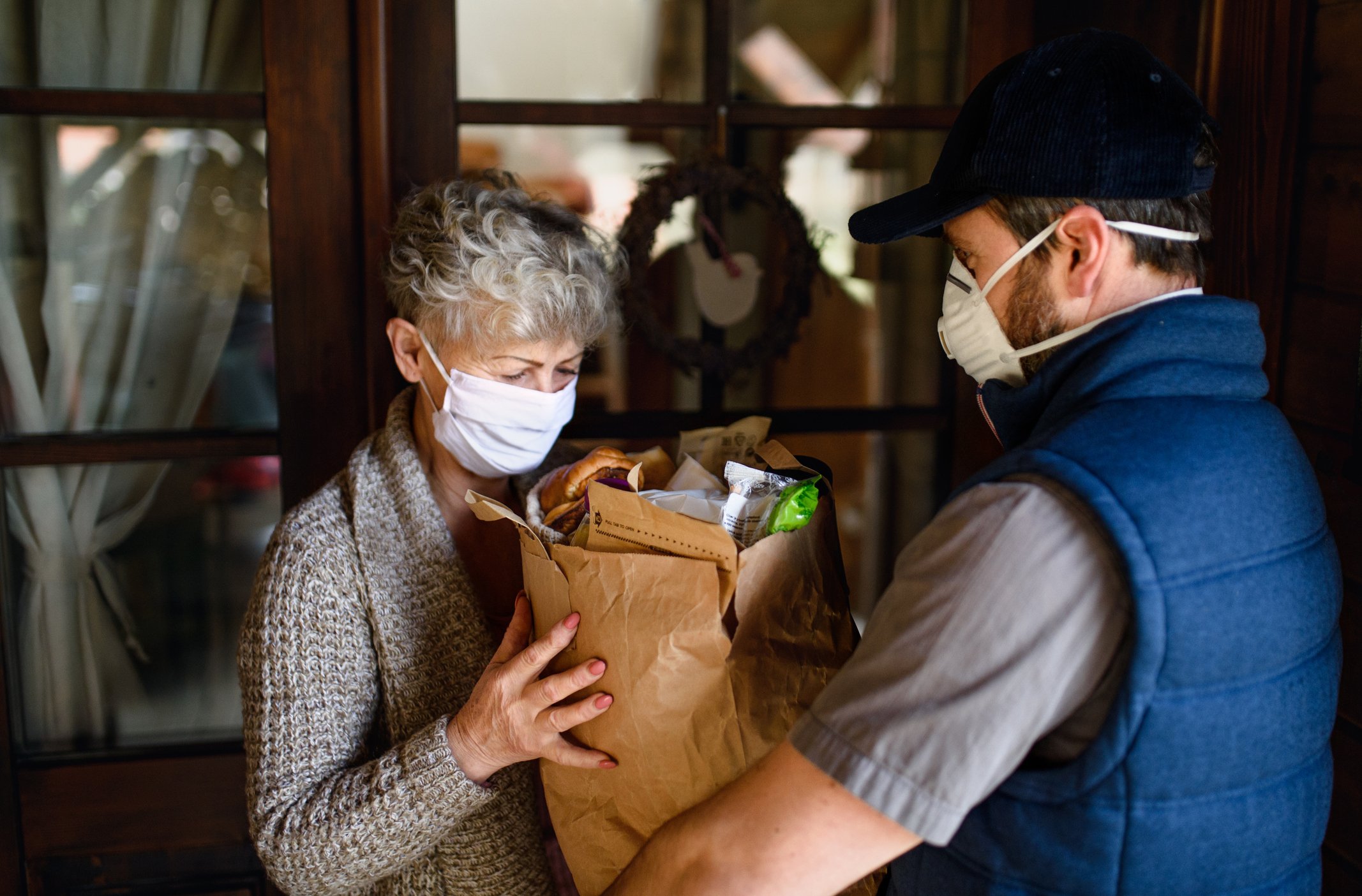 A delivery courier bringing groceries to an older person, both wearing face masks.