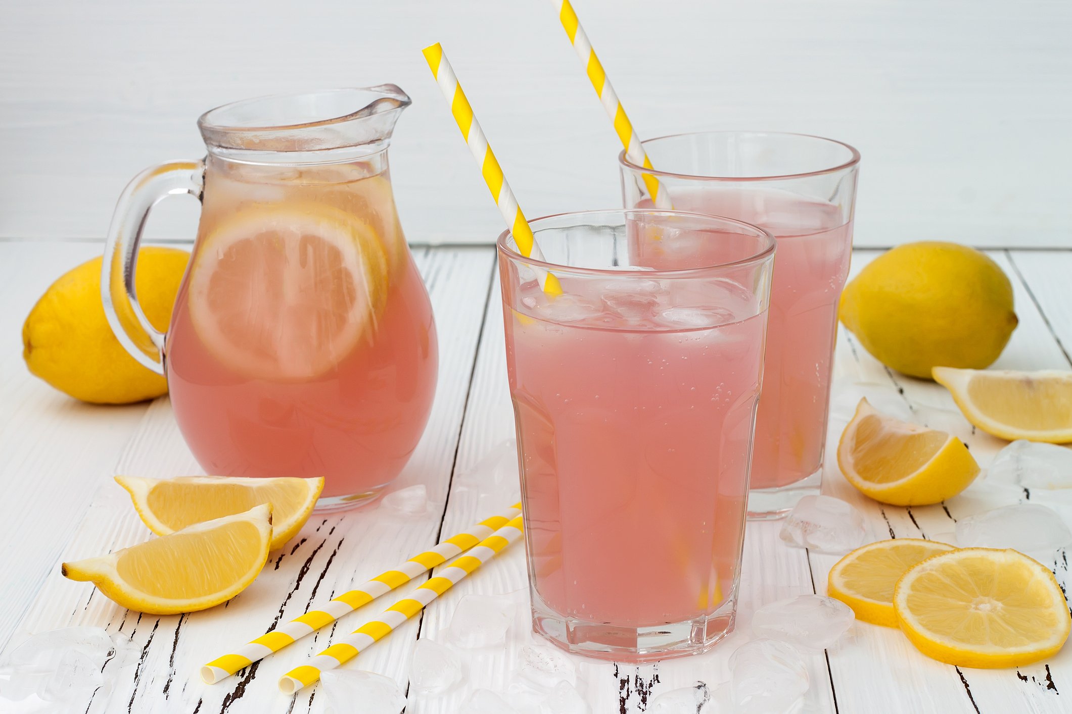 A pitcher and two cups of pink lemonade, surrounded by whole lemons and lemon wedges.
