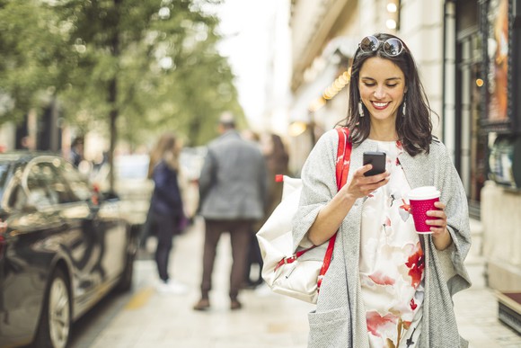A woman holding a coffee, looking at her phone, smiling as she walks down the street.