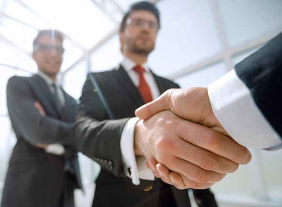 Two businesspeople shake hands as one watches approvingly in the background.