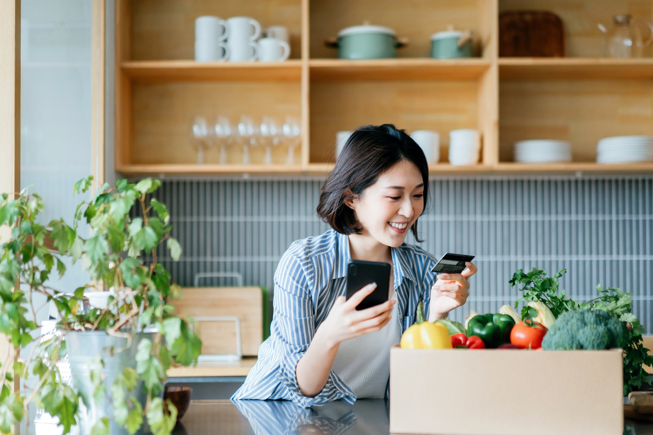 Woman on smartphone with credit card out leaning on a kitchen counter with a box of groceries. 