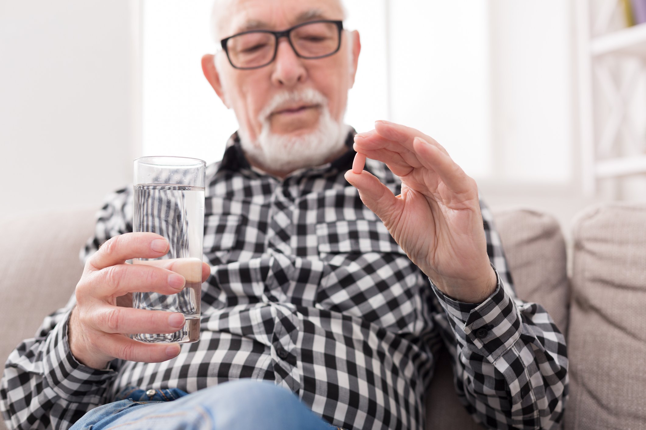 An elderly man wearing glasses and a flannel shirt reclining in a chair, holding a glass of water and a pill that he is about to ingest.