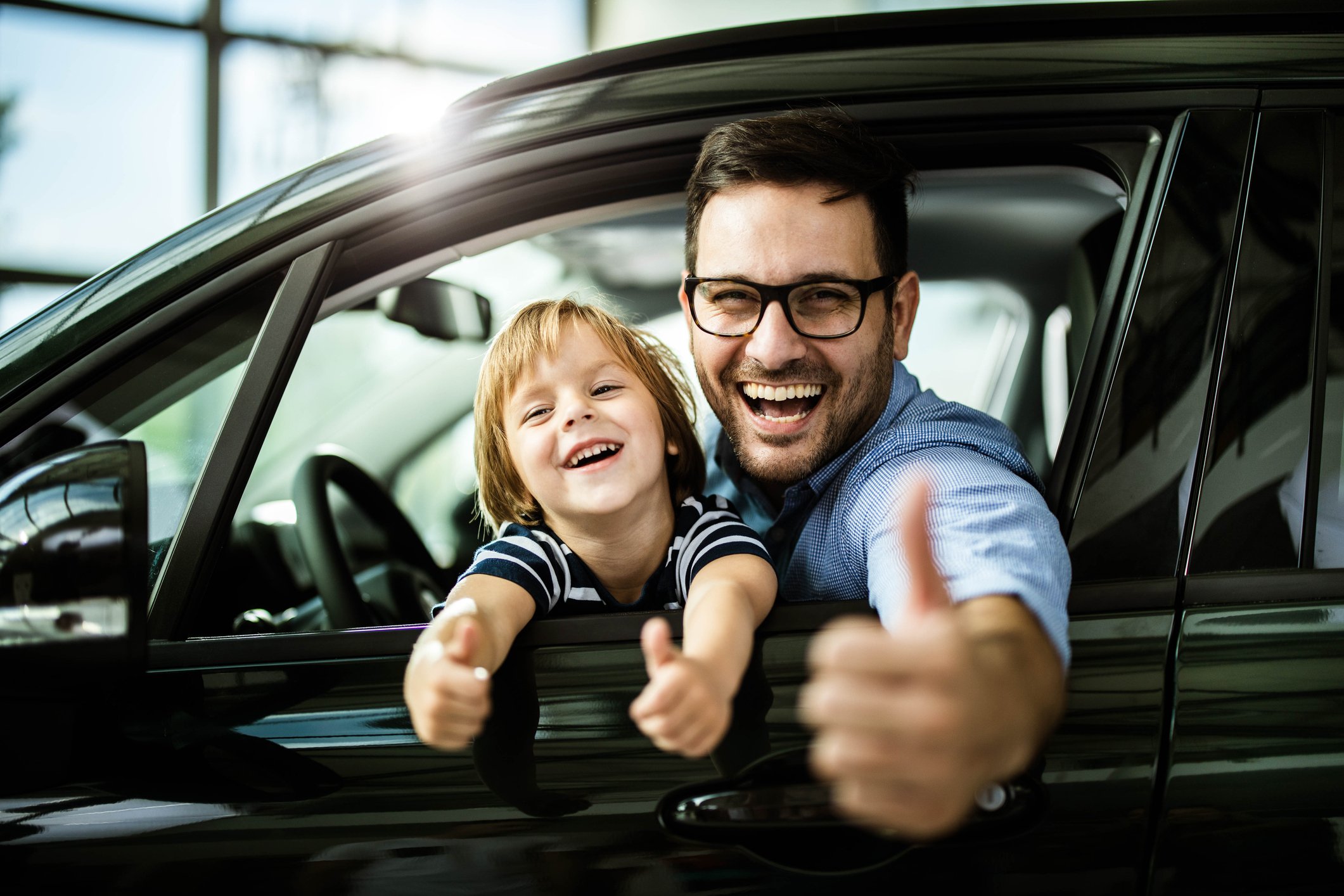 Parent and child in the driver's seat of a car, smiling and giving the thumbs-up.