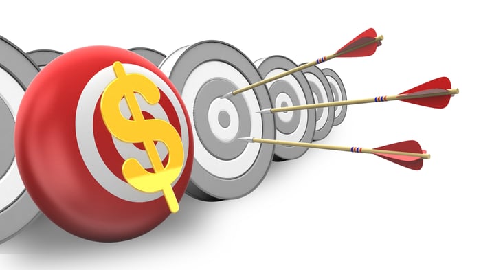 A red and white target with a yellow dollar on it next to several gray and white targets, one of which has three arrows stuck on it. 