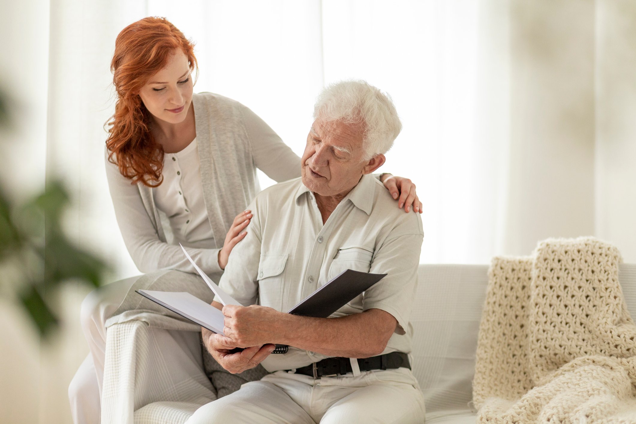 A young woman and an elderly man viewing a book of photos on a white couch.