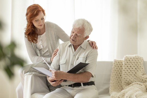 A young woman and an elderly man viewing a book of photos on a white couch.
