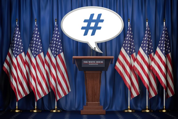 A speech bubble containing a hashtag is at a lectern with the words The White House labeling it and surrounded by American flags.