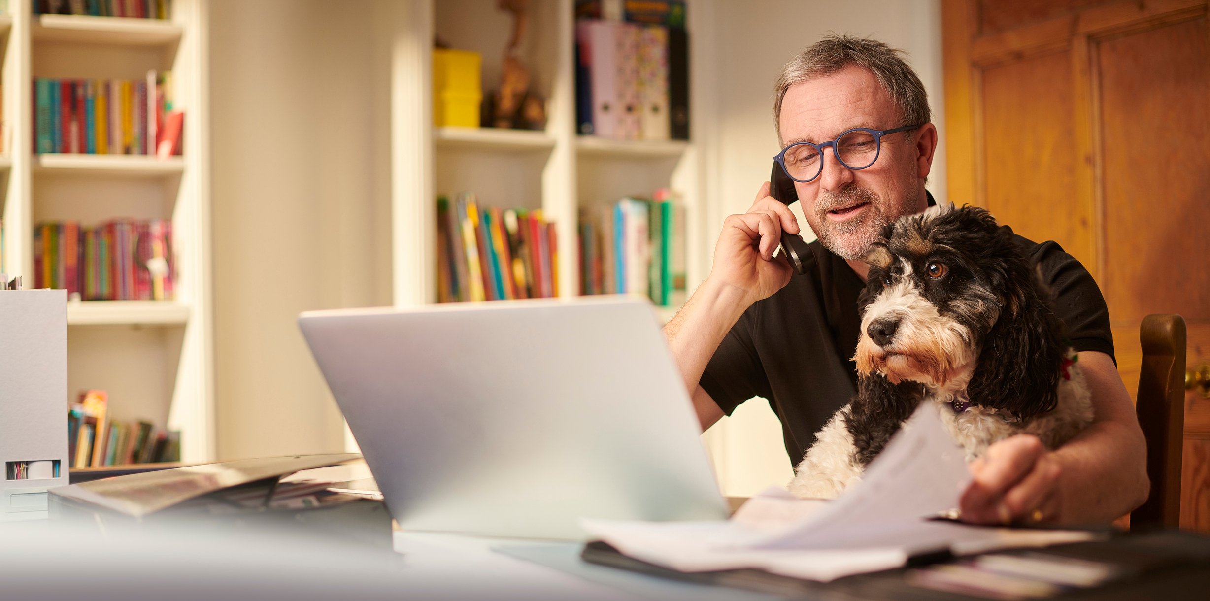 Man with dog on lap while looking at computer and talking on phone.