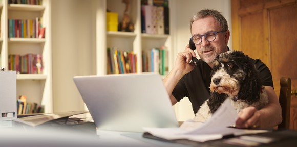 Man with dog on lap while looking at computer and talking on phone.