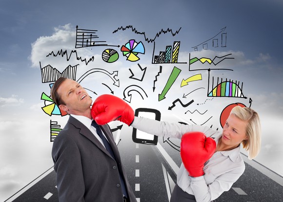 A businesswoman with boxing gloves landing a blow to a colleague with animated charts and graphs behind them.