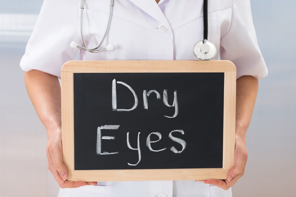 A person with a stethoscope around their neck holding a small chalkboard with "dry eyes" written on it