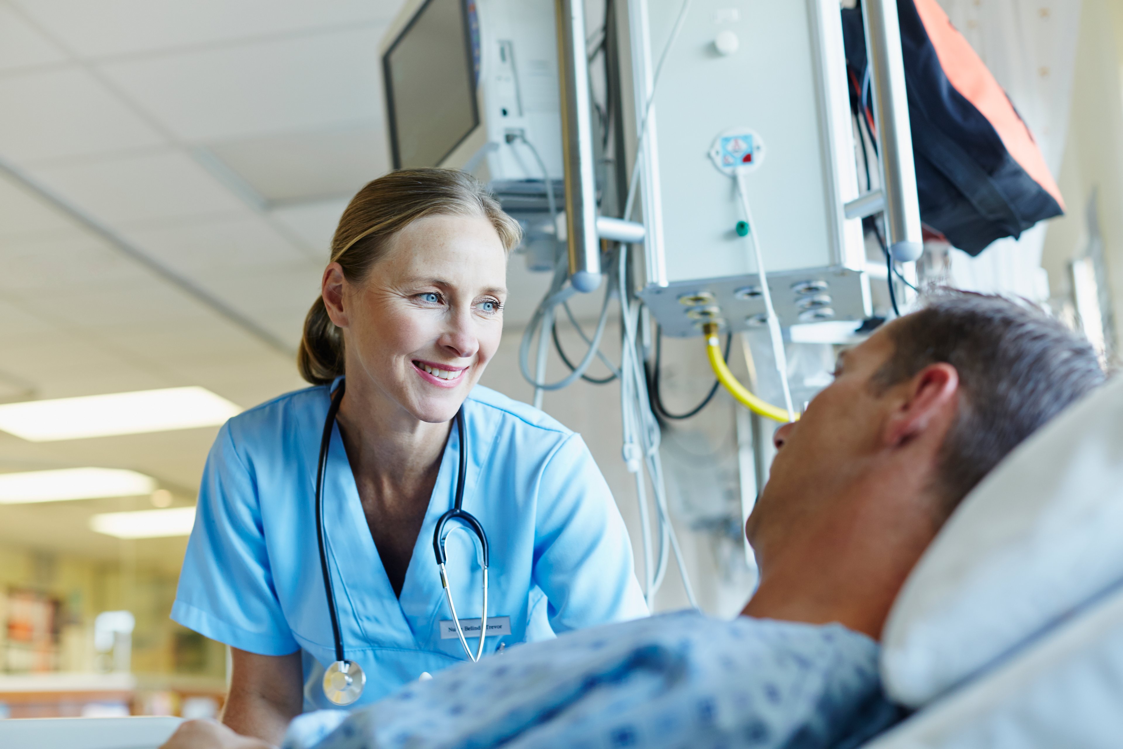 Female medical worker smiles at a patient laying in a hospital bed, medical devices in the hospital background.