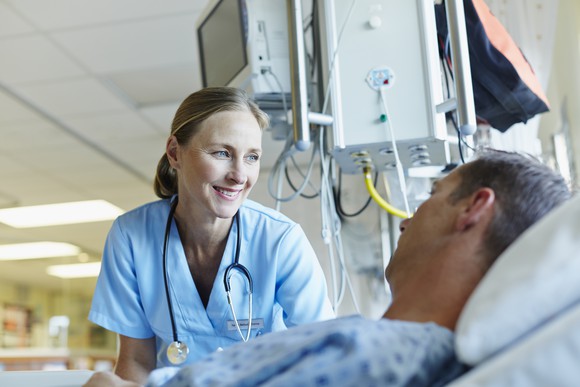 Female medical worker smiles at a patient laying in a hospital bed, medical devices in the hospital background.
