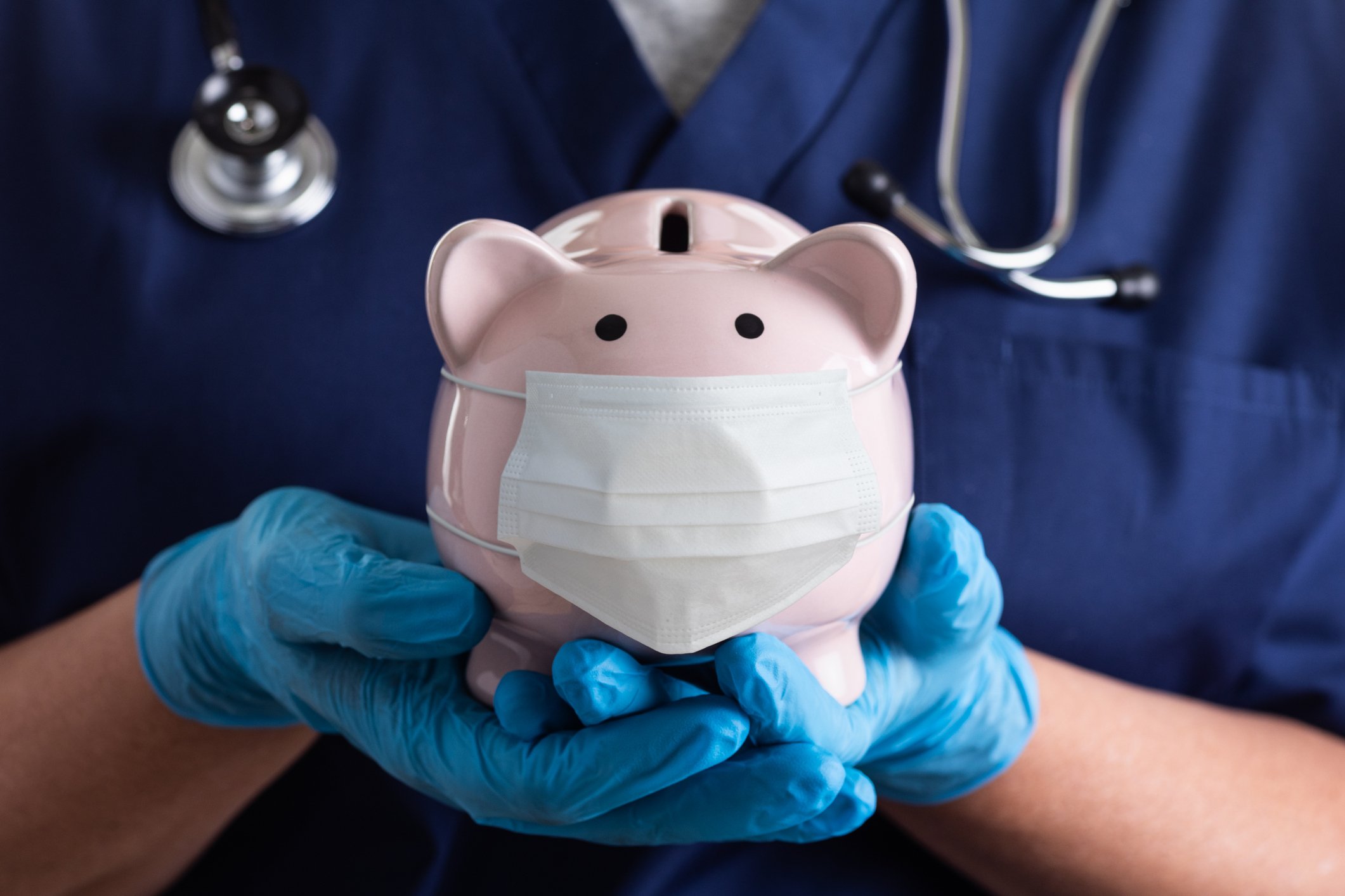 A doctor holds a piggy bank that's wearing a surgical mask.