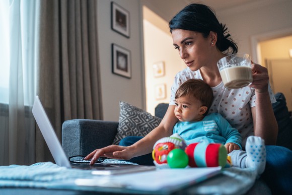 A young mother using a laptop while holding a measuring cup while her toddler stares at the computer screen.